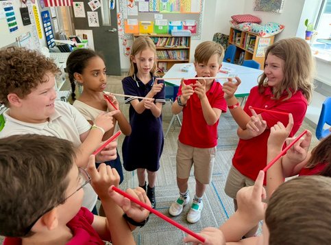 A circle of Selwyn students each balancing a red rod on their fingertips together during class.